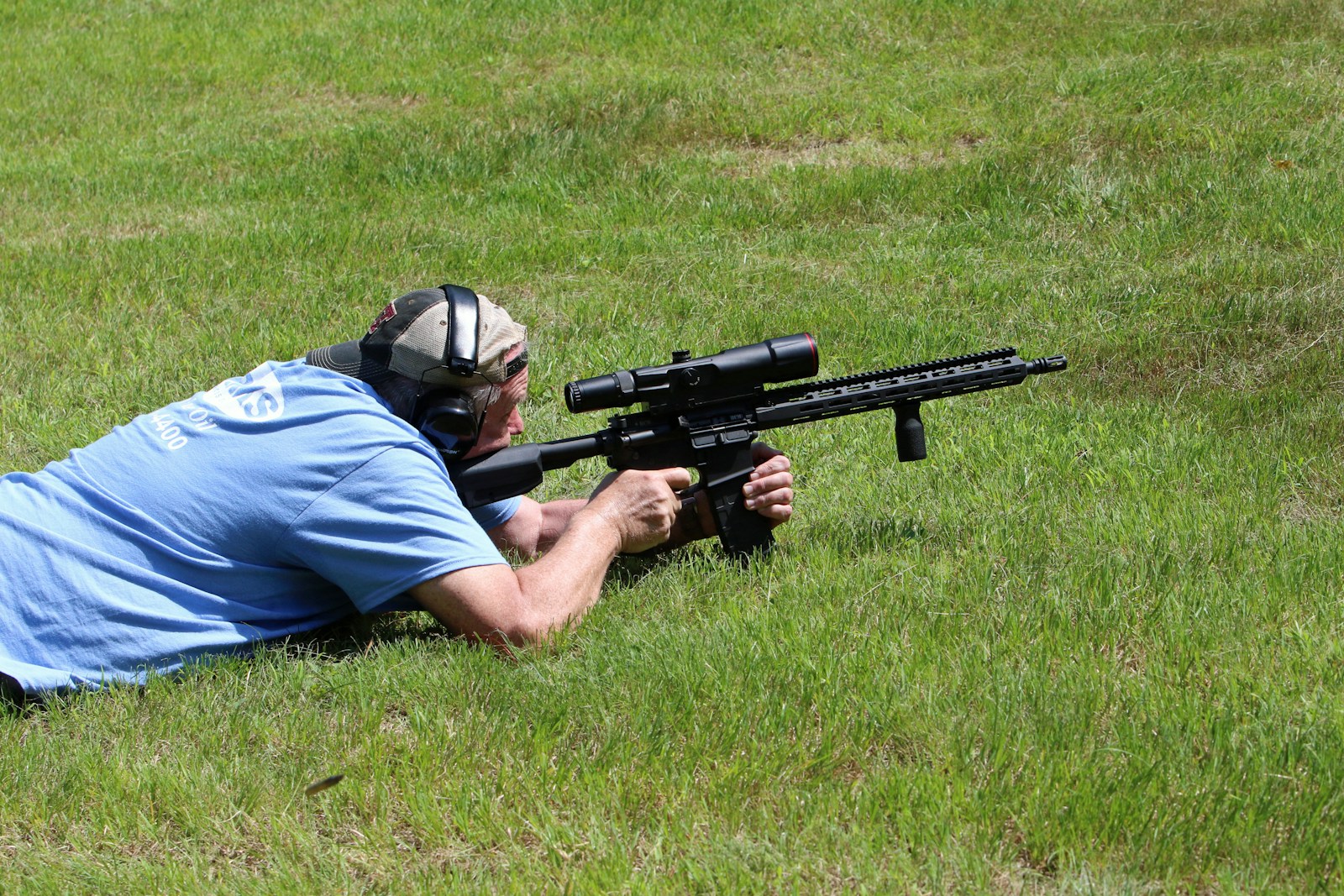 Man lies prone, aiming a rifle on grass.