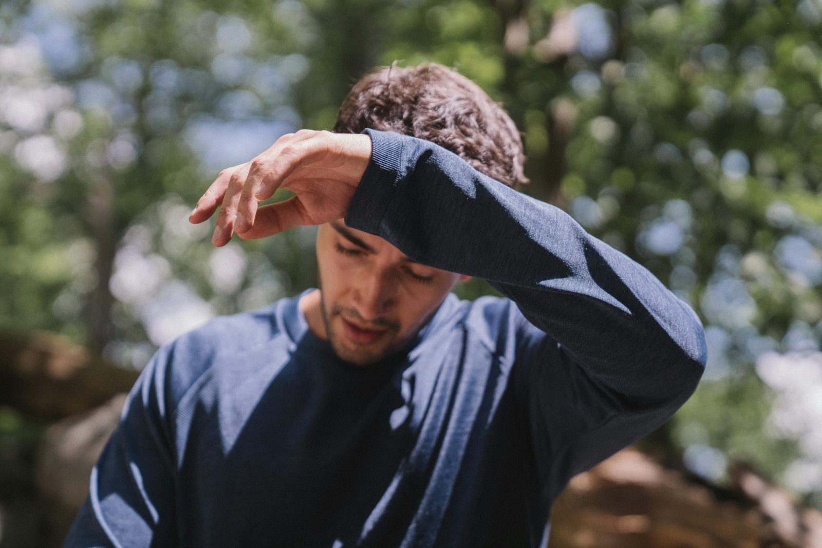 An adult man outdoors with eyes closed, appears pensive among lush green foliage.
