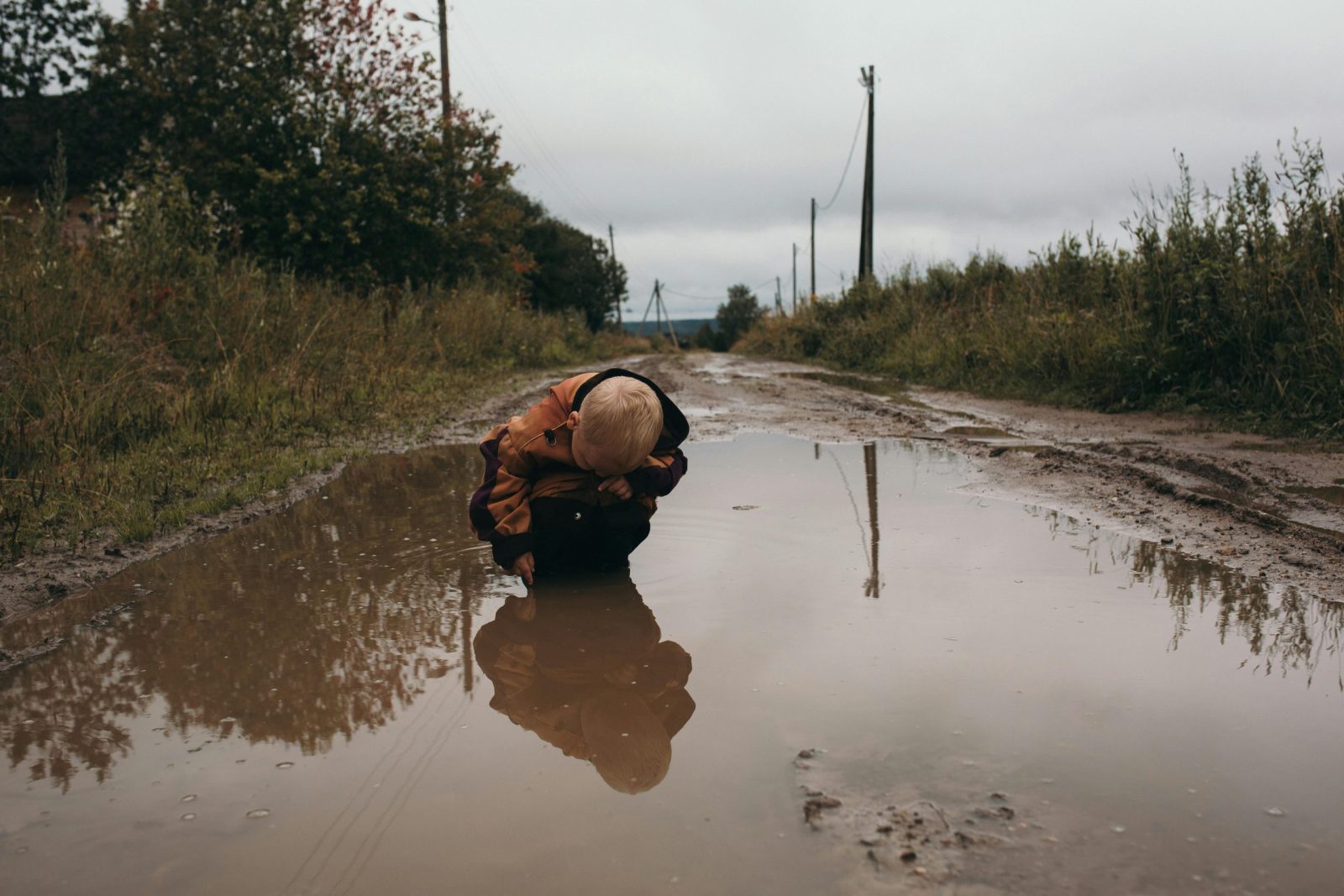 A young child fascinated by a puddle on a muddy rural road, exploring nature.