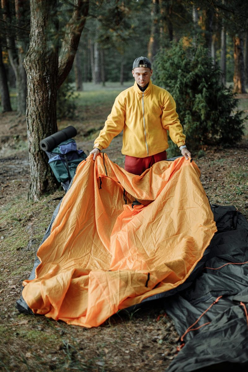 A young man sets up a bright orange tent in a peaceful forest campsite, preparing for an adventure.