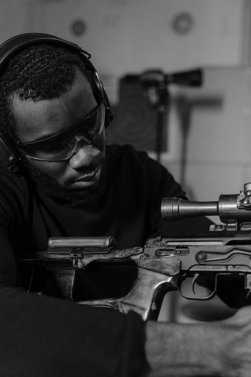 Black and white photo of a man adjusting a rifle scope indoors.