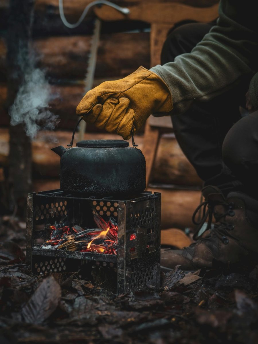 Close-up of a kettle on a camping stove with glove-covered hand adjusting it outdoors.