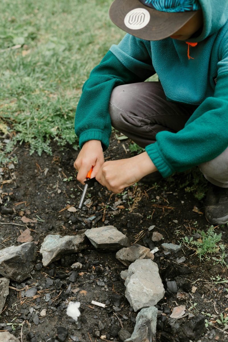 Boy in green hoodie using fire starter outdoors on rocky ground in Russia.