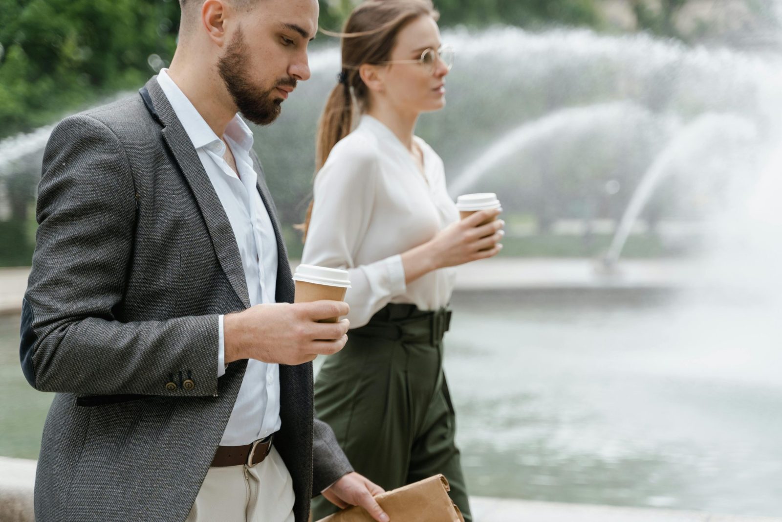 Two professionals enjoying a coffee walk through a city park, showcasing formal attire and togetherness.