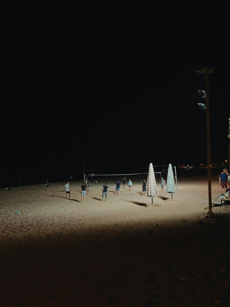 A group of people play volleyball on a sandy beach at night, illuminated by lights near umbrellas.