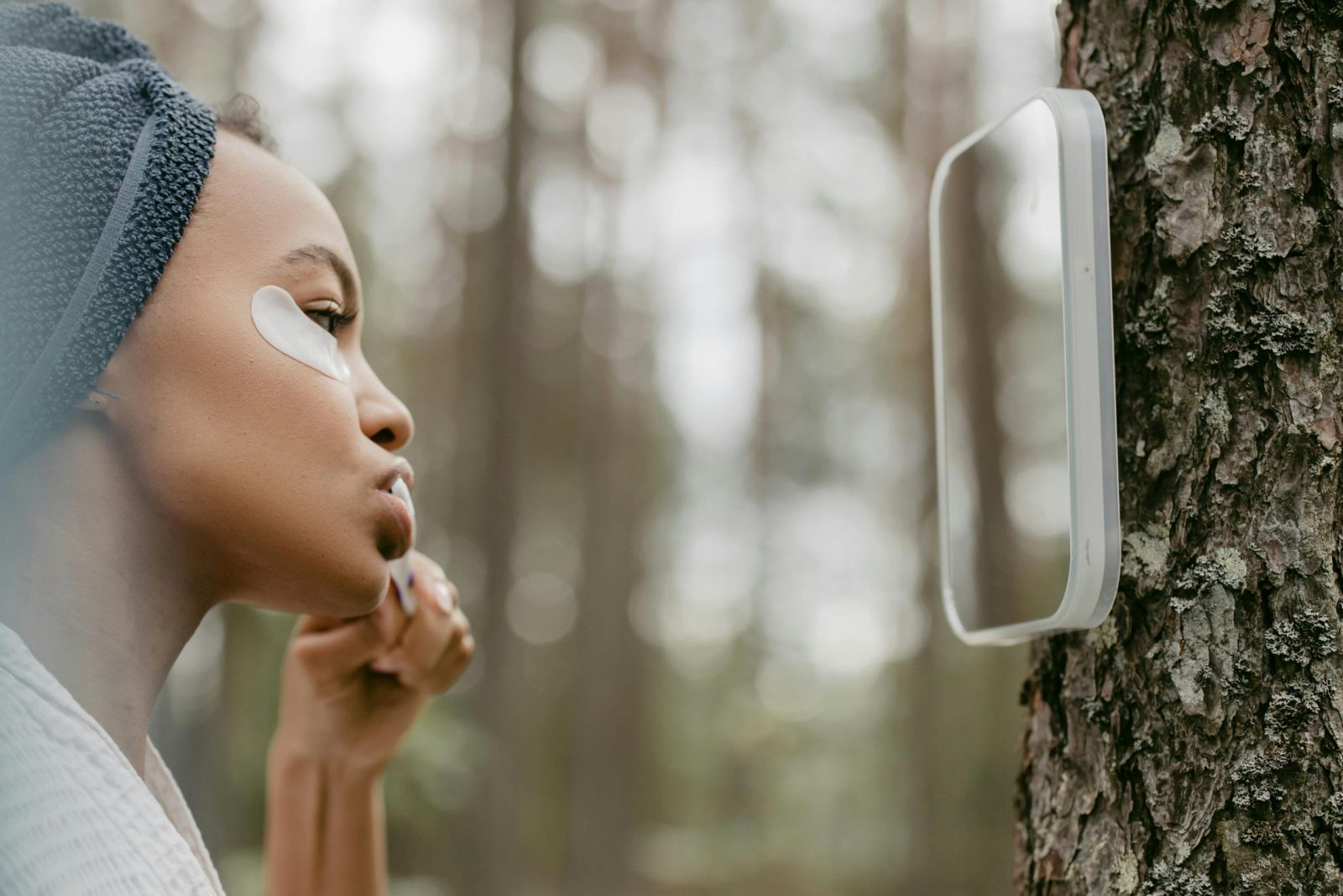 Side view of a woman with an eye patch brushing teeth in a forest using a mirror.