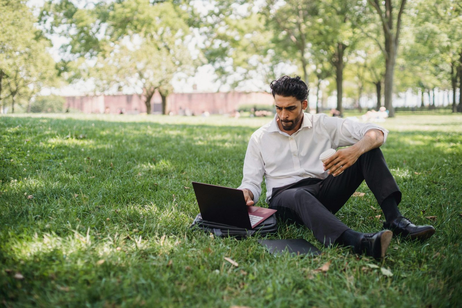 Businessman sitting on grass in park, working on laptop and holding coffee.