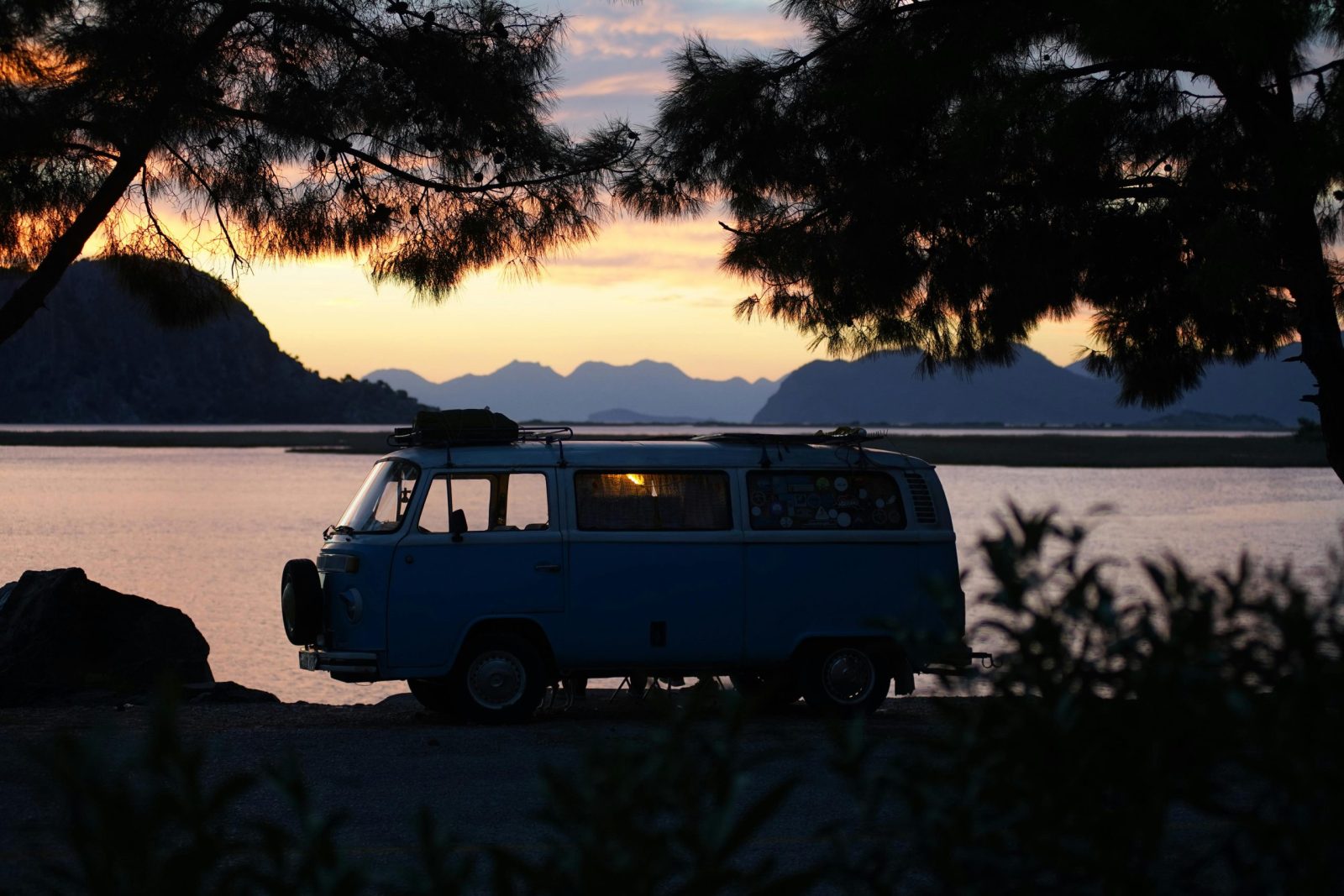 A vintage van parked by a calm lakeside, silhouetted against the sunset, surrounded by trees and mountains.