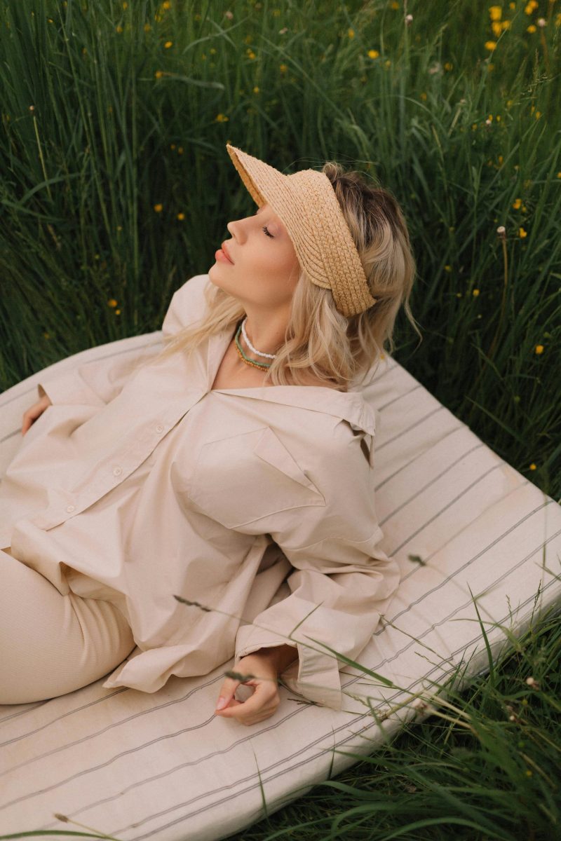 Woman in casual attire relaxing on a mattress outside in a grassy area, wearing a hat and basking in nature.
