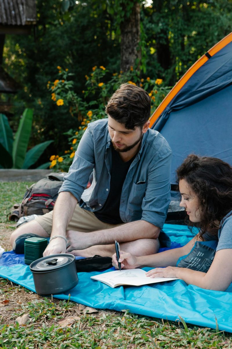 Calm young female camper lying on blanket and drawing in notebook near boyfriend during picnic in green woods on sunny day