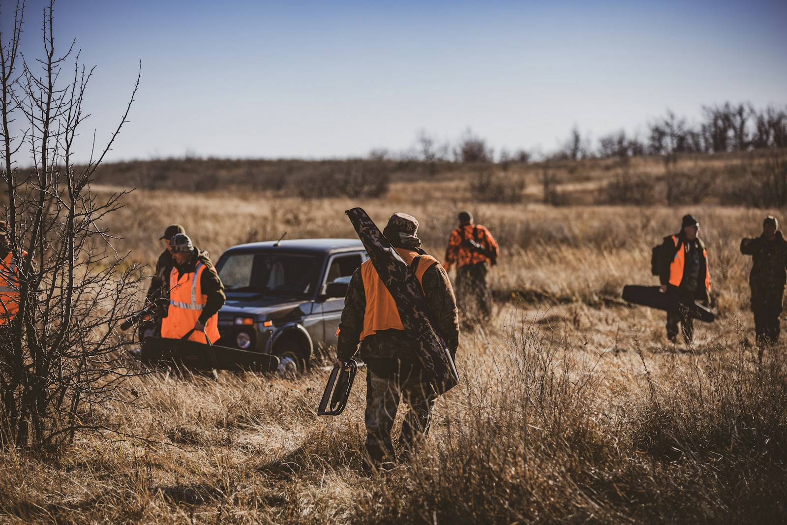 Hunters walk through dry grass near a vehicle.