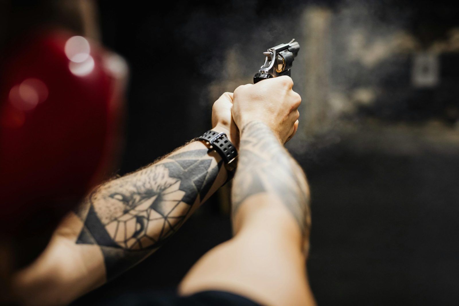 Close-up of tattooed arms aiming and firing a revolver in an indoor shooting range.