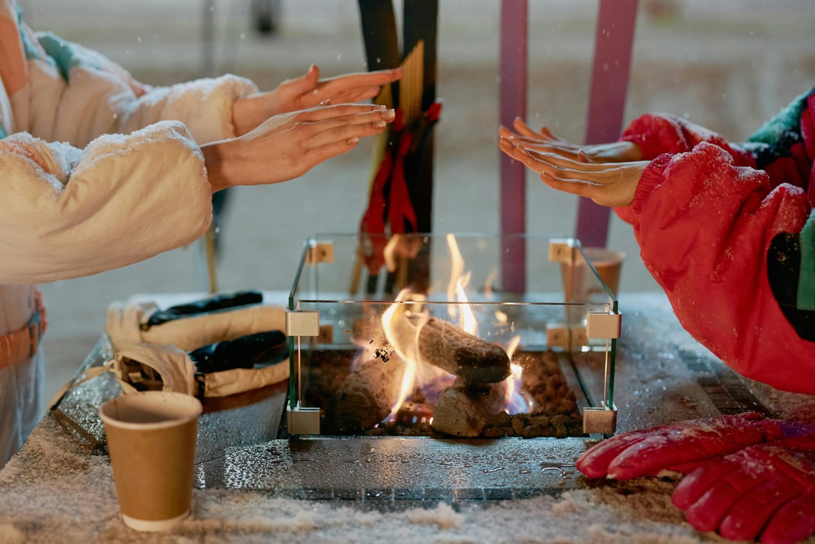 Two people warming hands over a glass-enclosed outdoor fire on a snowy day.
