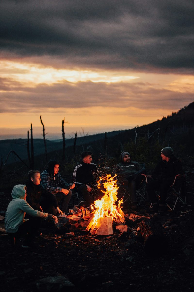A group of friends sitting around a campfire in a mountainous area at sunset, enjoying the outdoors.