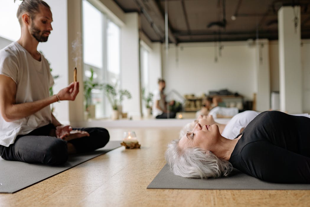 Yoga Instructor Sitting Cross-legged with Incense Over Elderly Women Lying on Mats