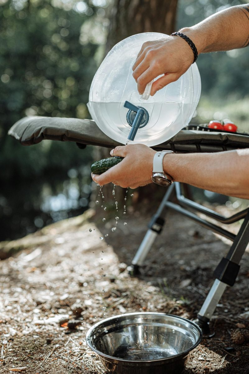 Person washing hands with portable water dispenser and bowl outdoors, promoting hygiene in nature.