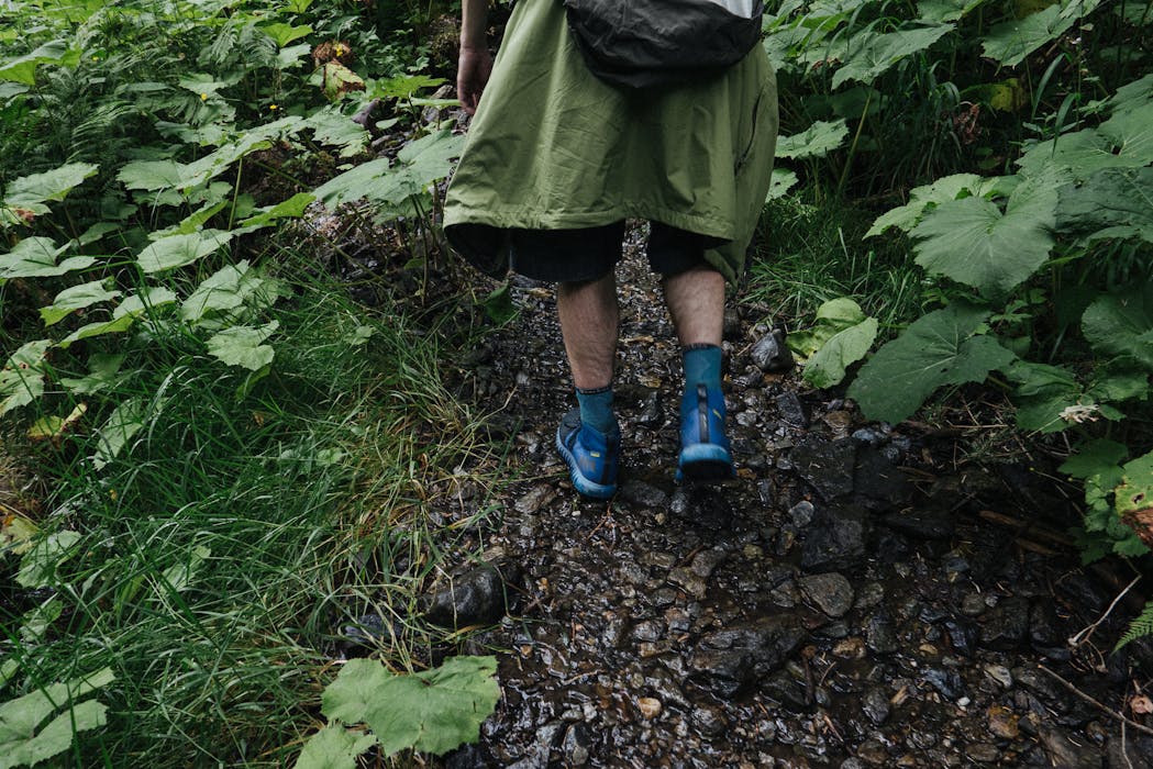 A Person in Green Jacket and Blue Rubber Shoes Standing on Green Grass Field