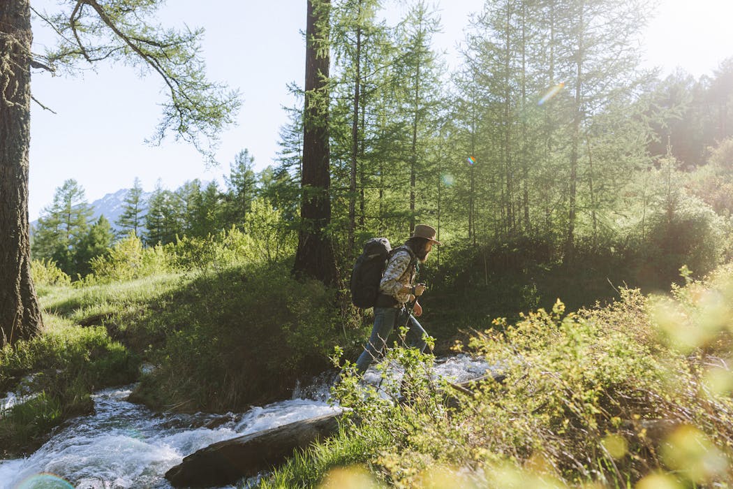 Man in Brown and White Long Sleeve Shirt Carrying Black Backpack Walking on Brown Tree Log Near River