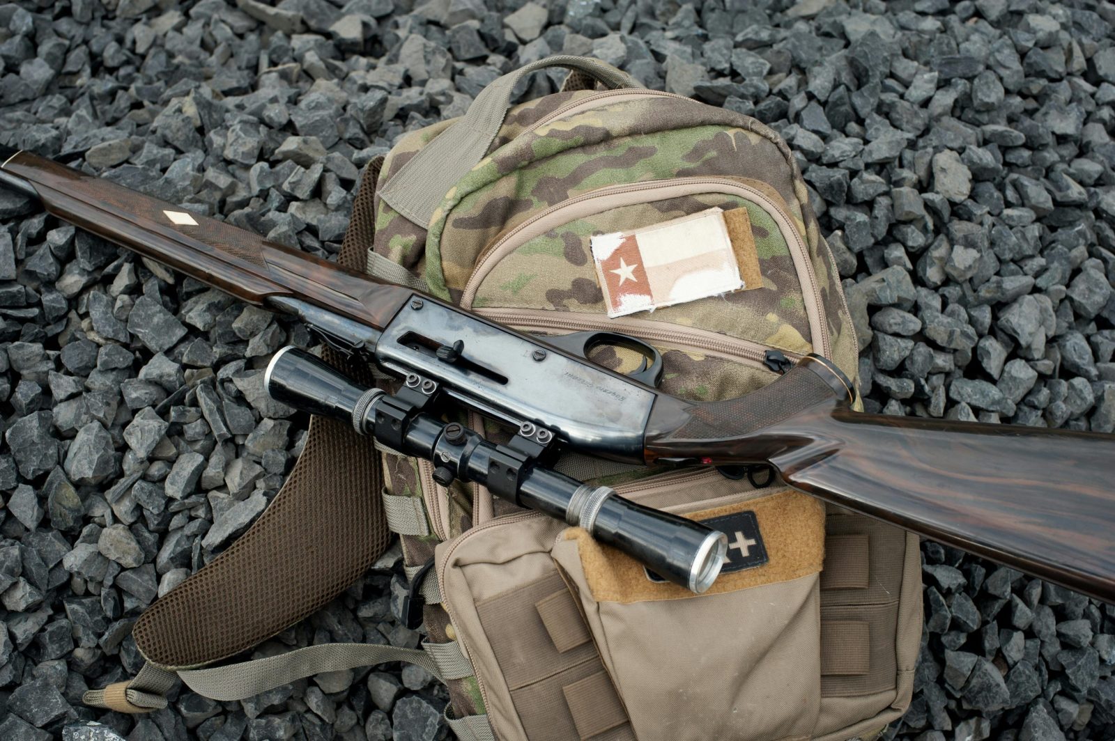 Camouflage backpack with sniper rifle on pebbles, symbolizing military preparedness.