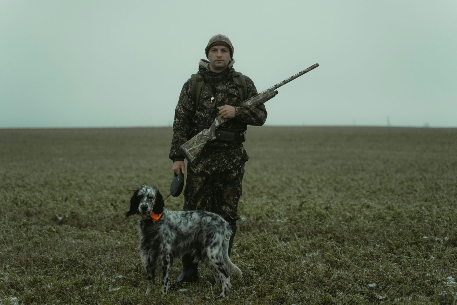 A hunter in camouflage gear stands with a dog in an expansive field, showcasing outdoor adventure.