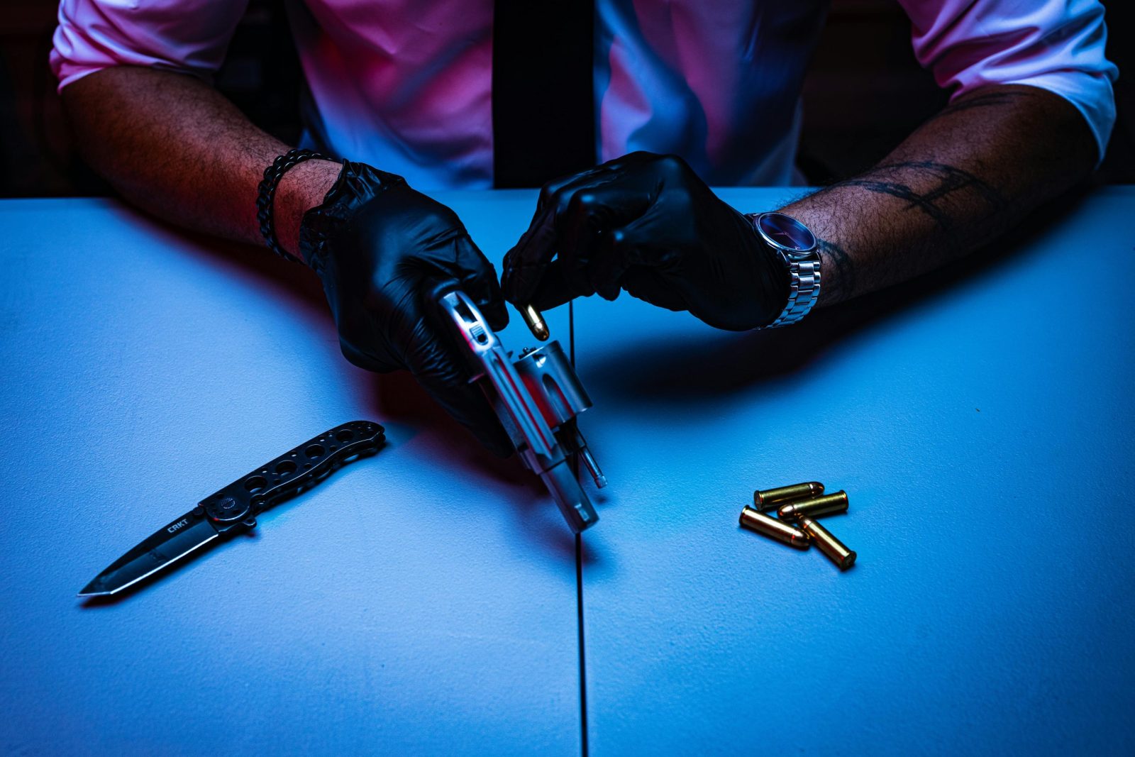 Man with gloves handling a revolver and knife on a table with bullets nearby.