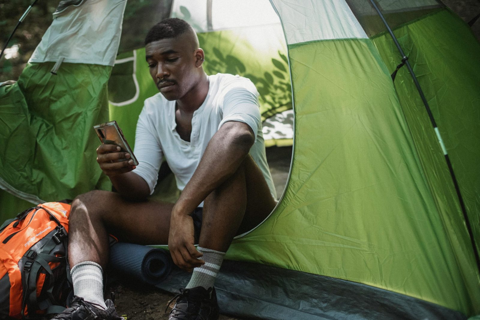 Sad African American male traveler resting in tent and browsing Internet on cellphone while spending journey in nature