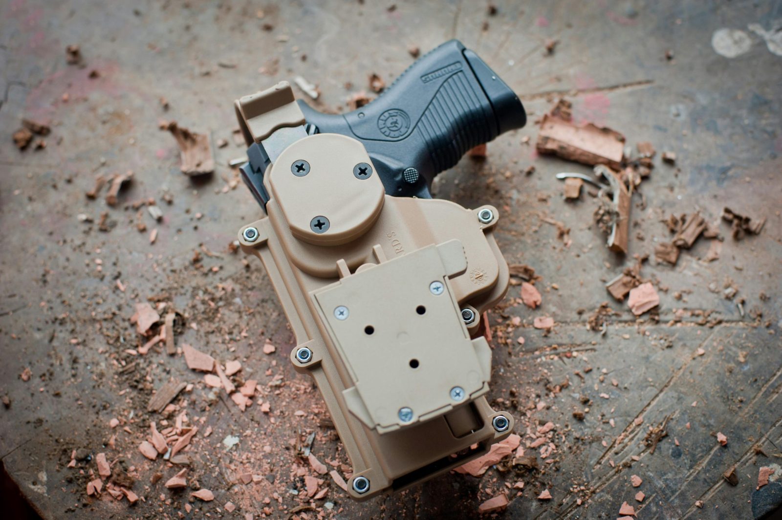 A close-up view of a pistol secured in a tactical holster, placed on a dusty and rough concrete surface with scattered debris.