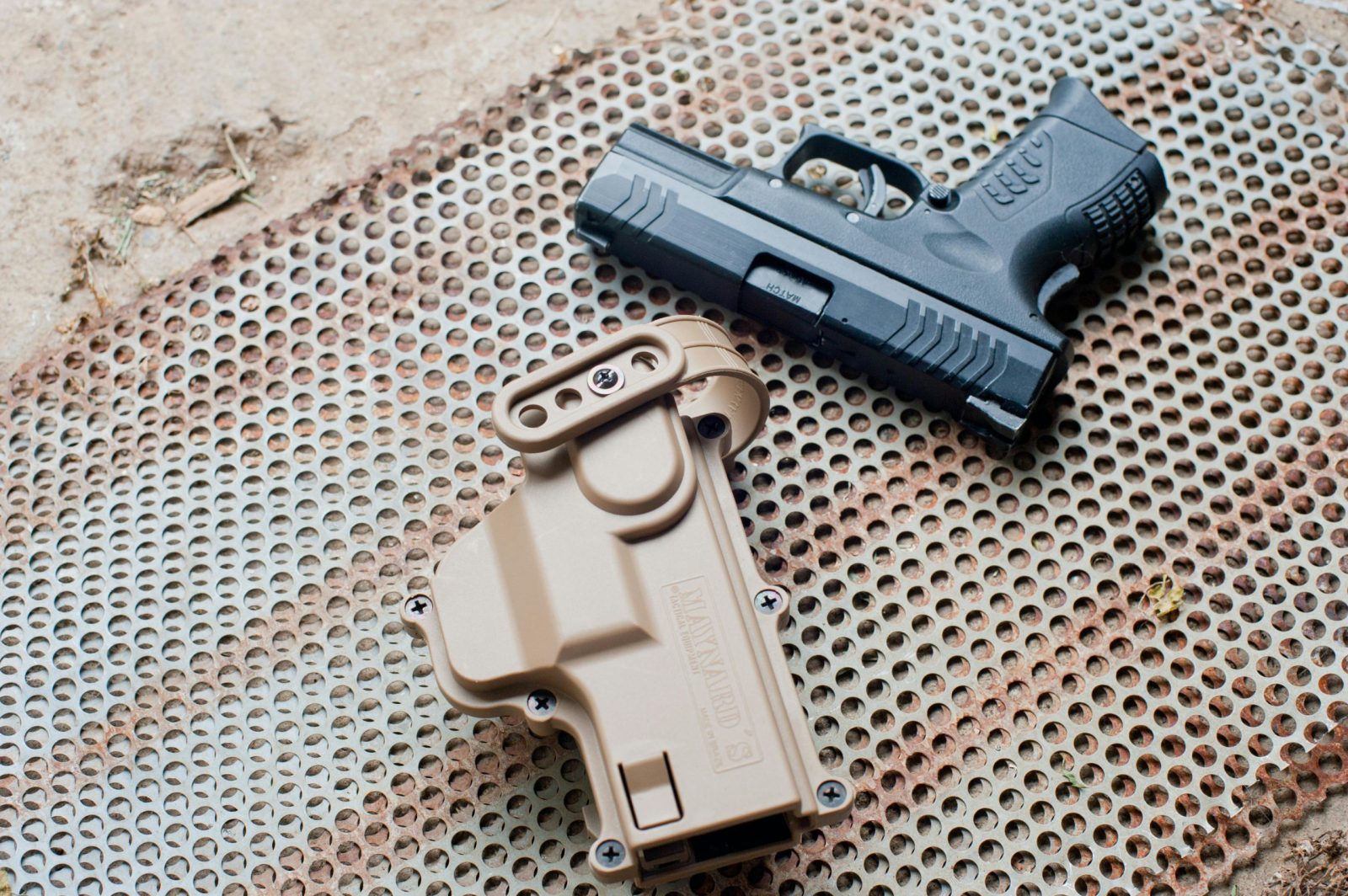 Detailed top view of a black pistol beside a beige holster on a perforated metal surface outdoors.