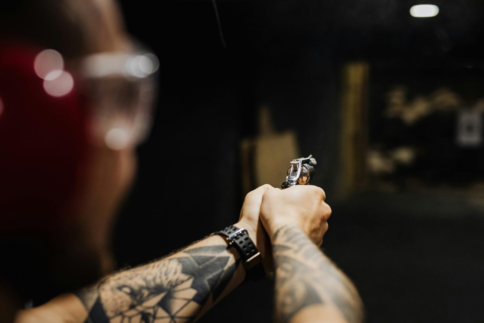 Man with arm tattoo aims a handgun at an indoor shooting range, wearing protective gear.