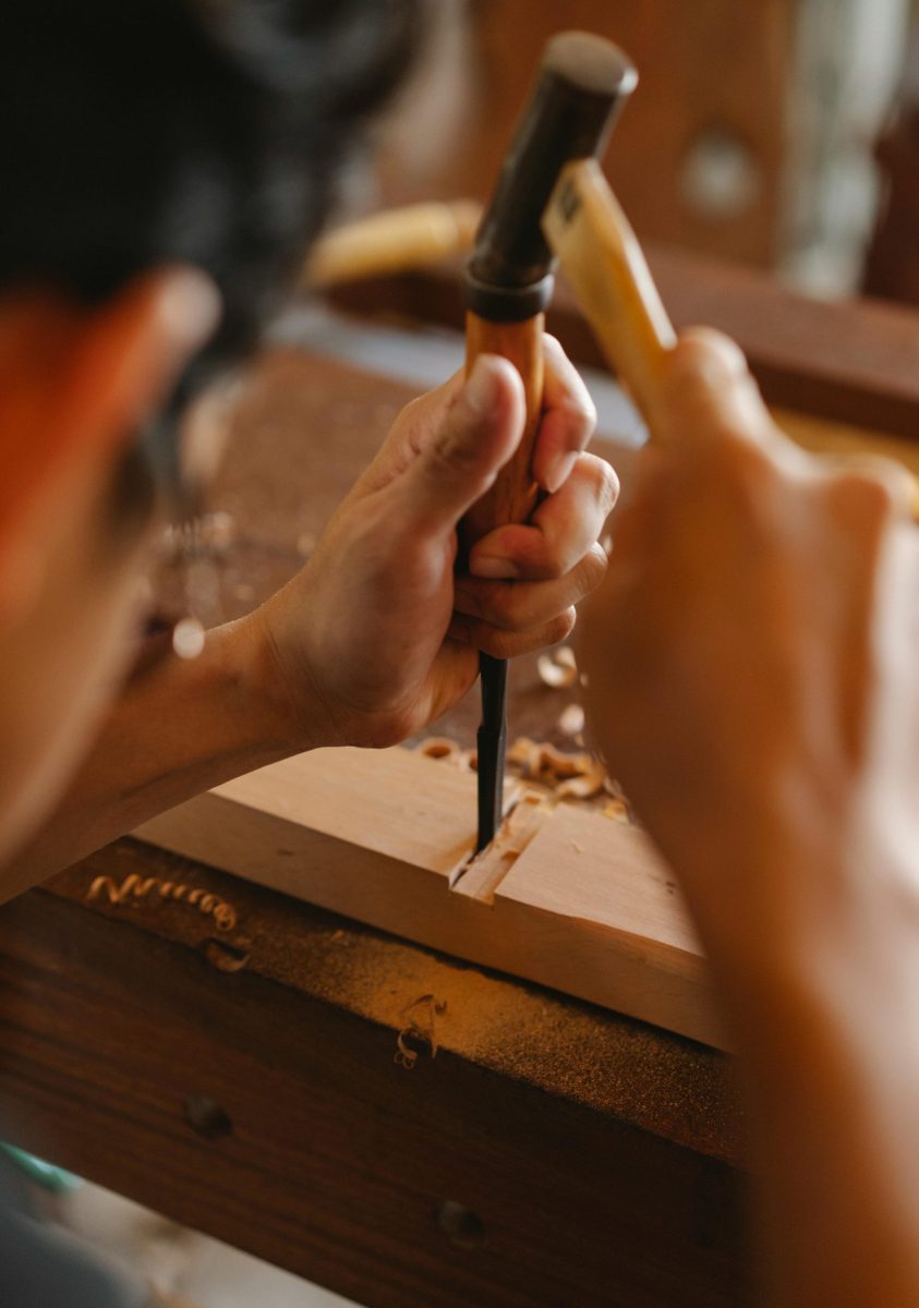 Crop focused artisan making notch in hardwood plank using chisel and small hammer