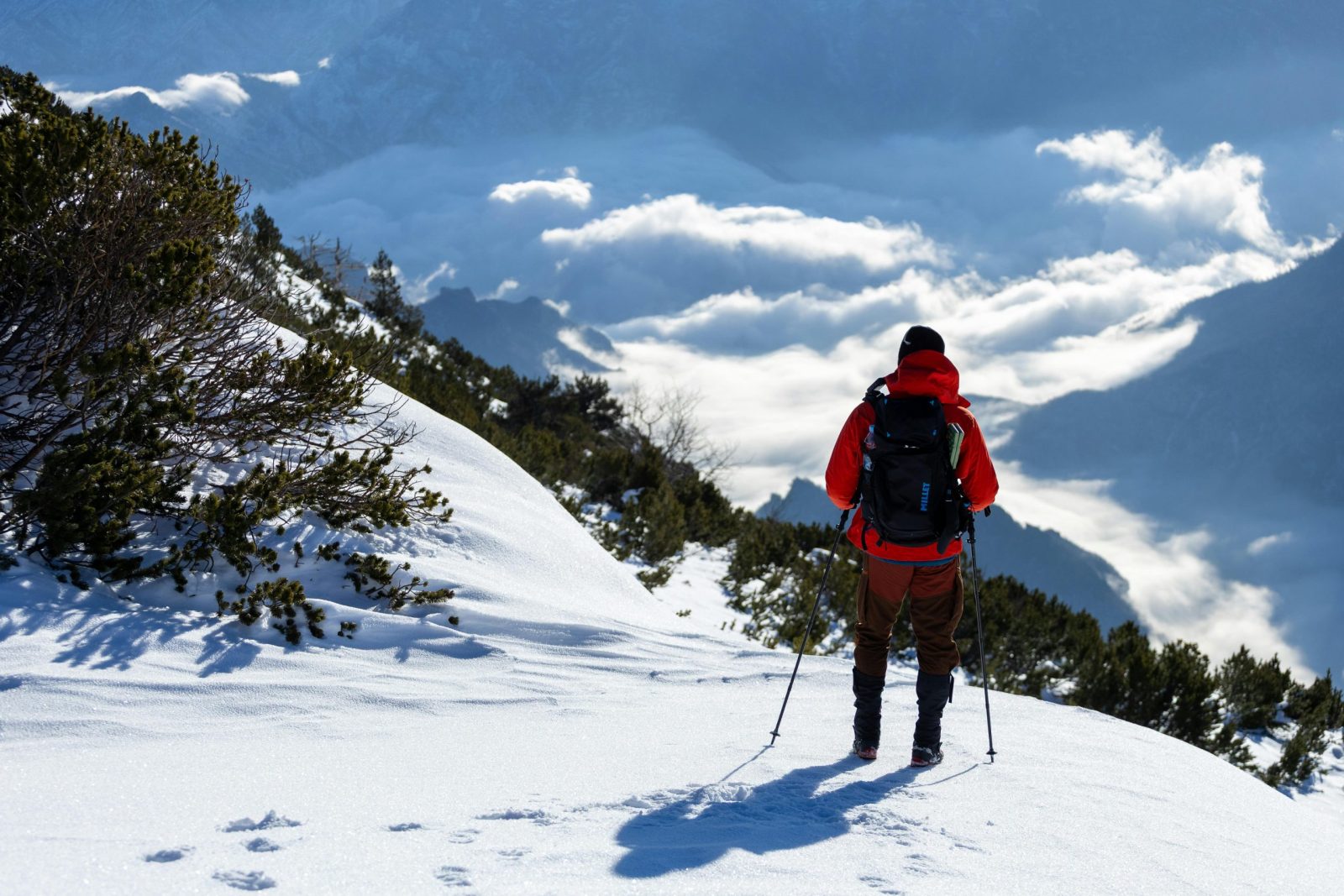 A lone hiker in red gear traverses a snow-covered mountain slope with a stunning view of clouds below.