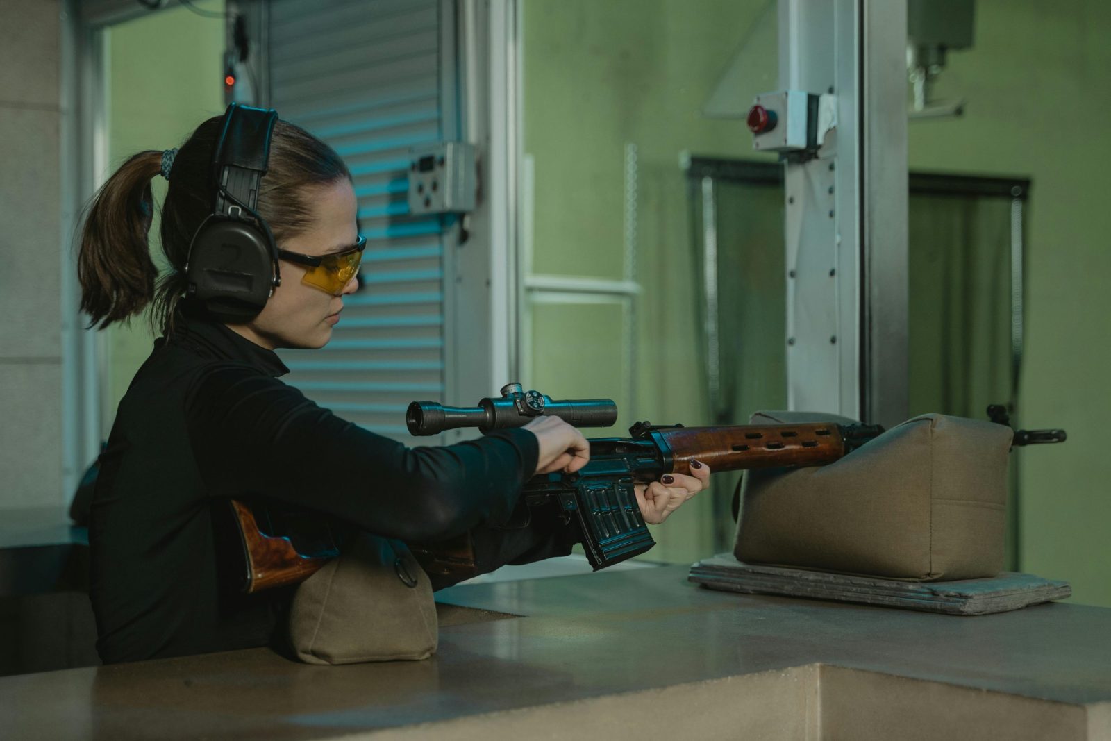 Focused woman aiming a sniper rifle at an indoor shooting range wearing ear and eye protection.