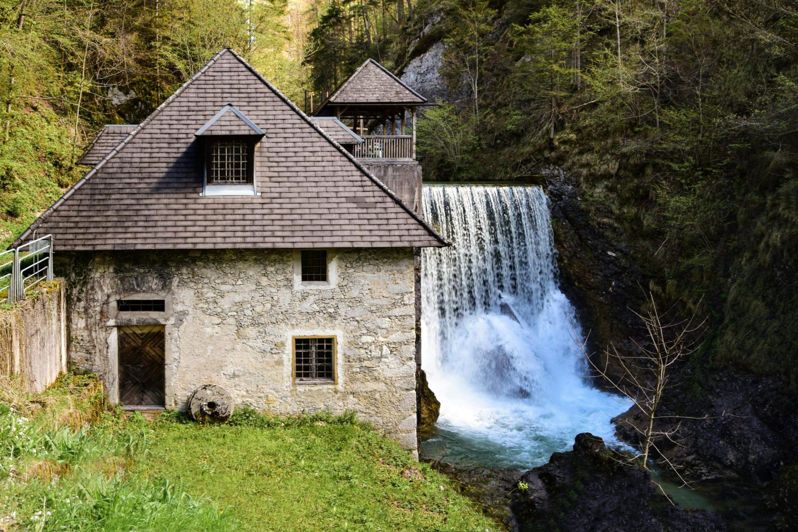 Charming rustic stone house beside a picturesque waterfall in a lush forest setting.
