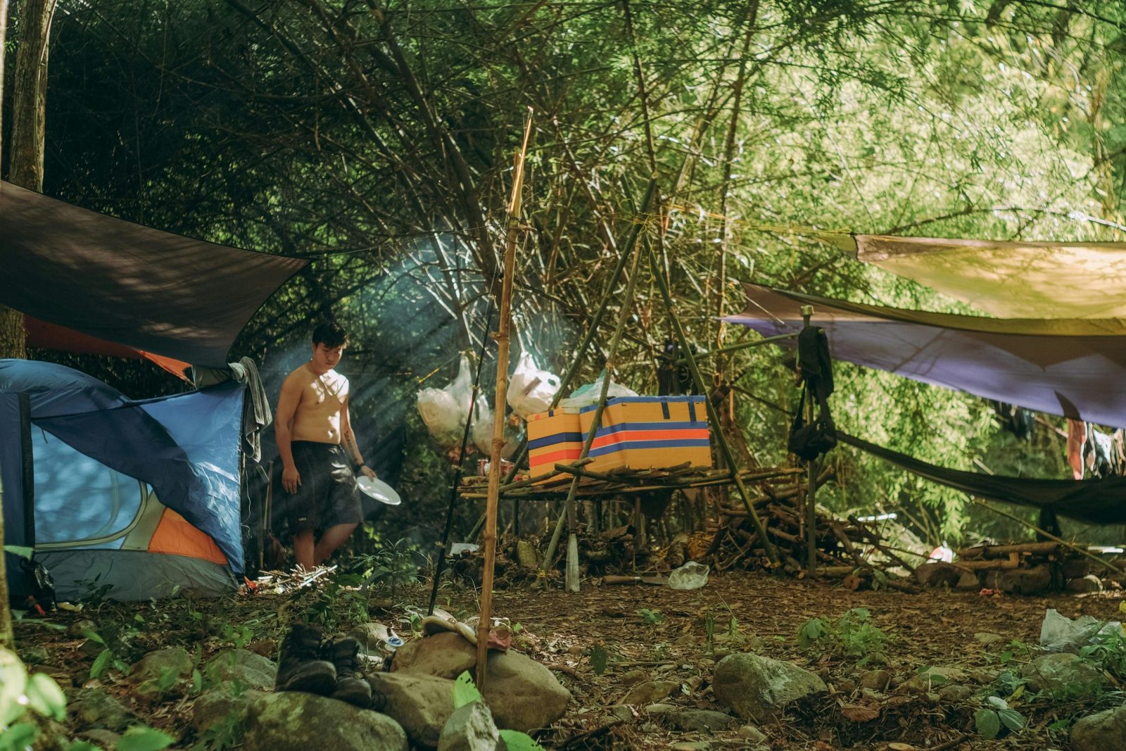 A peaceful forest campsite featuring tents, hammocks, and a shirtless man in natural daylight.