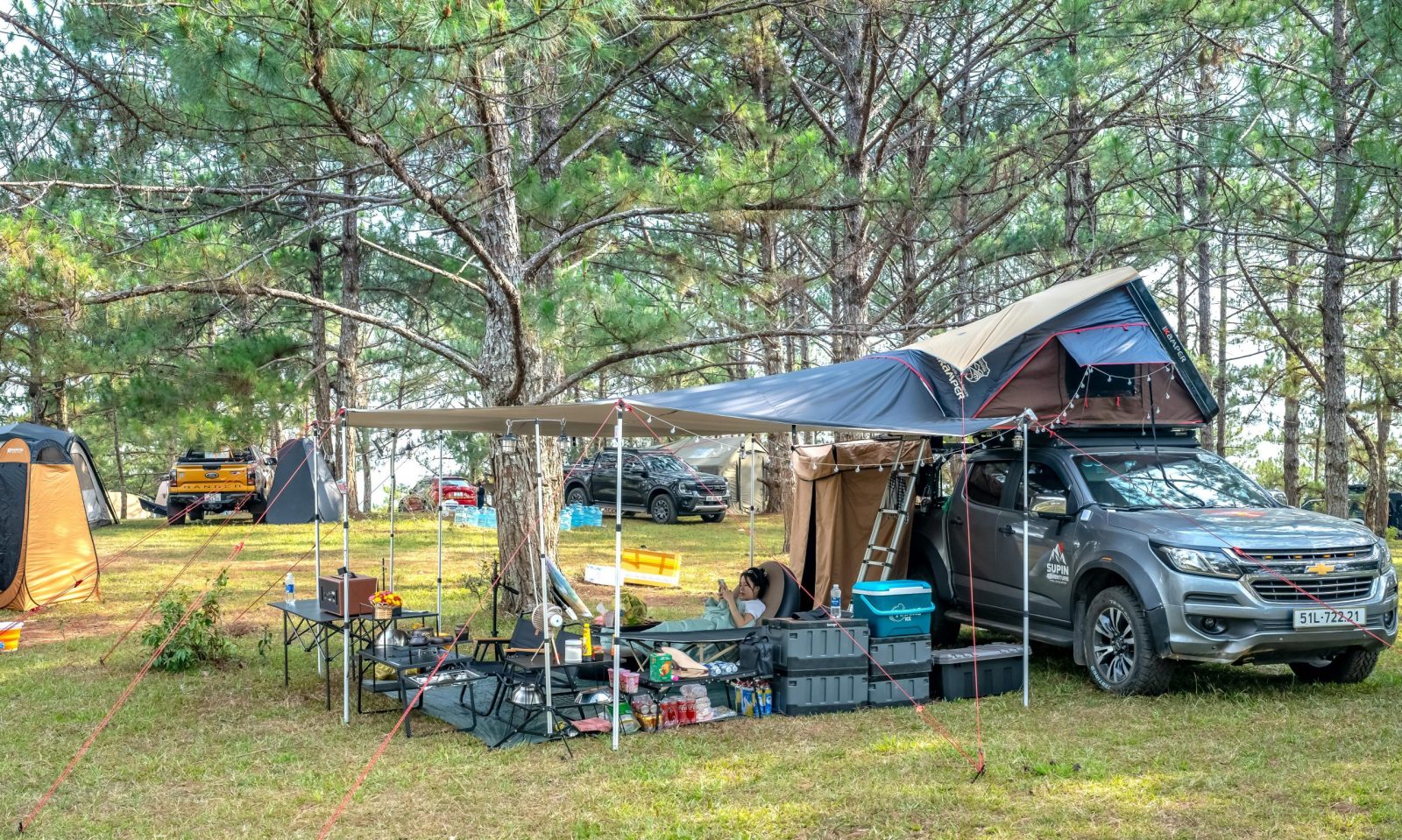 Outdoor camping scene featuring cars, tents, and a rooftop tent in a wooded area.