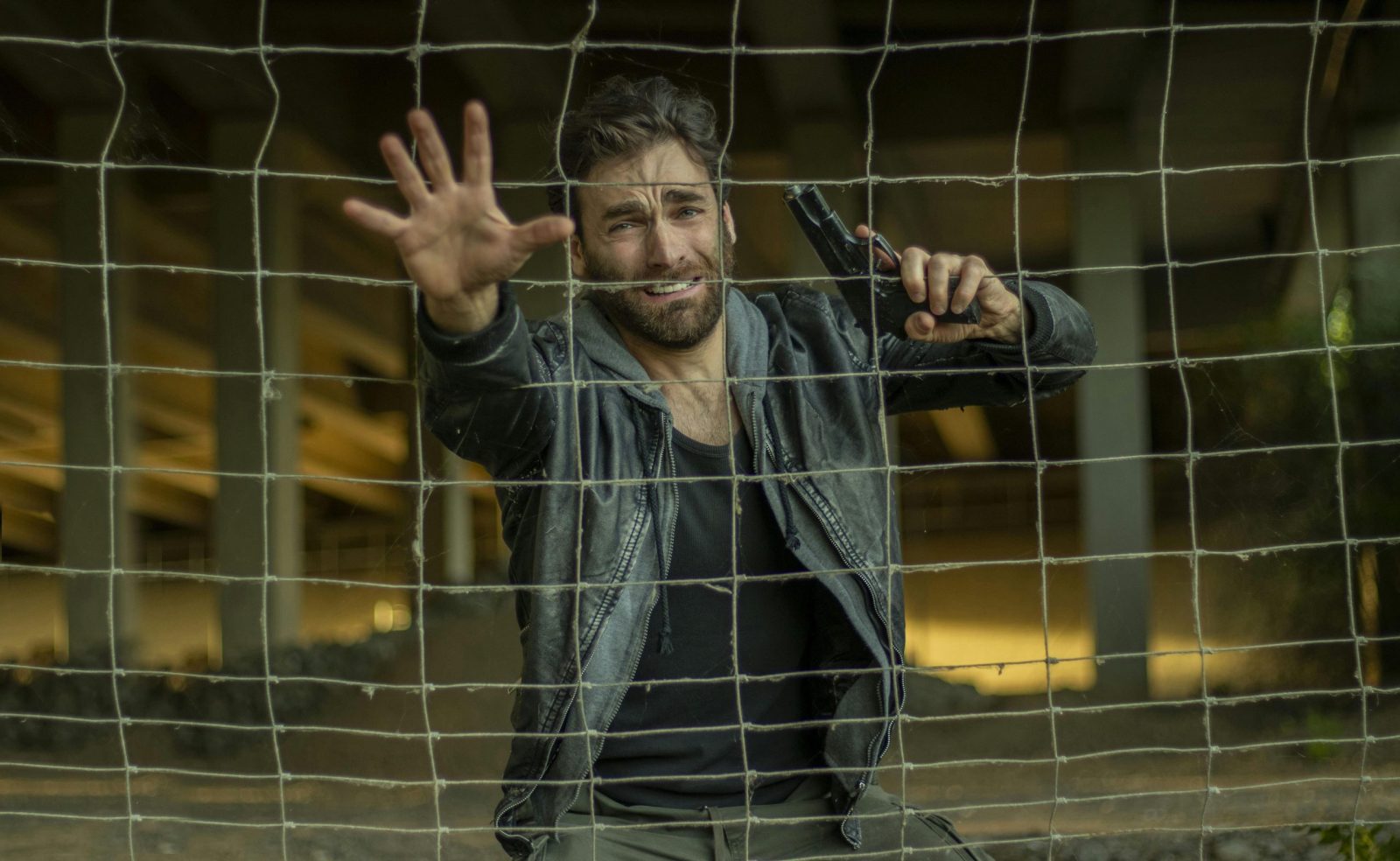 A distressed man reaches through a fence, holding a gun, in a tense setting.