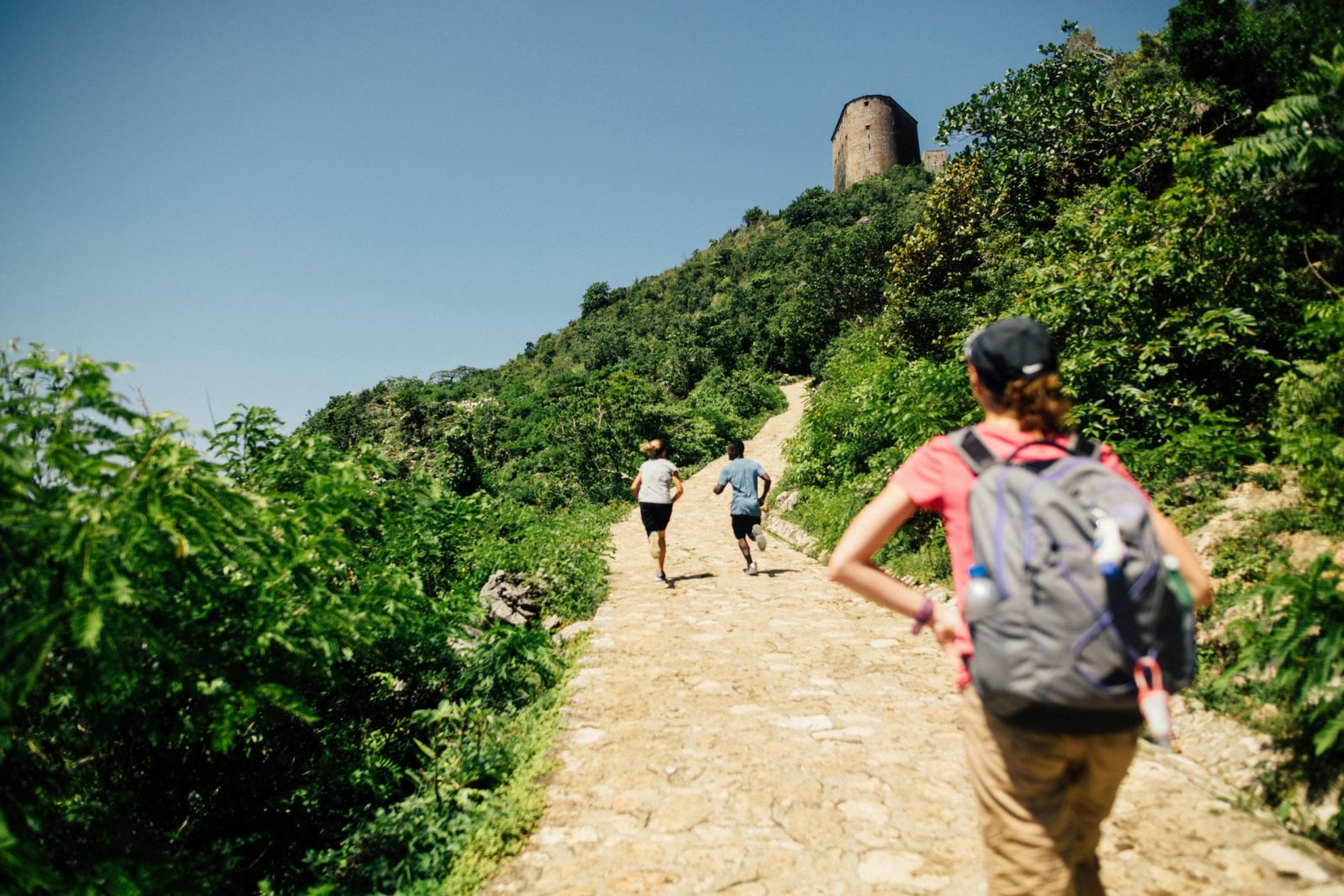People hiking on a sunny mountain trail with a tower in the background, enjoying nature and leisure.