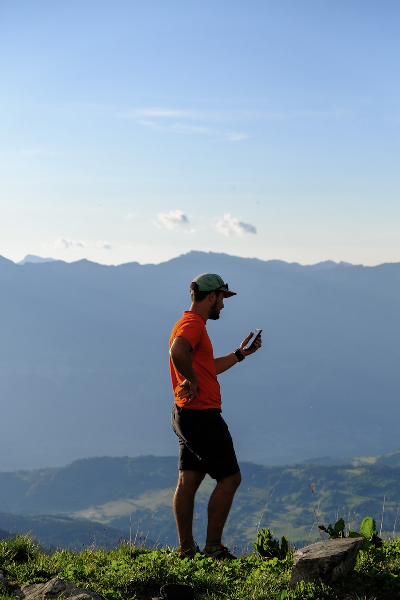 man in orange tank top and black shorts holding camera standing on top of mountain during